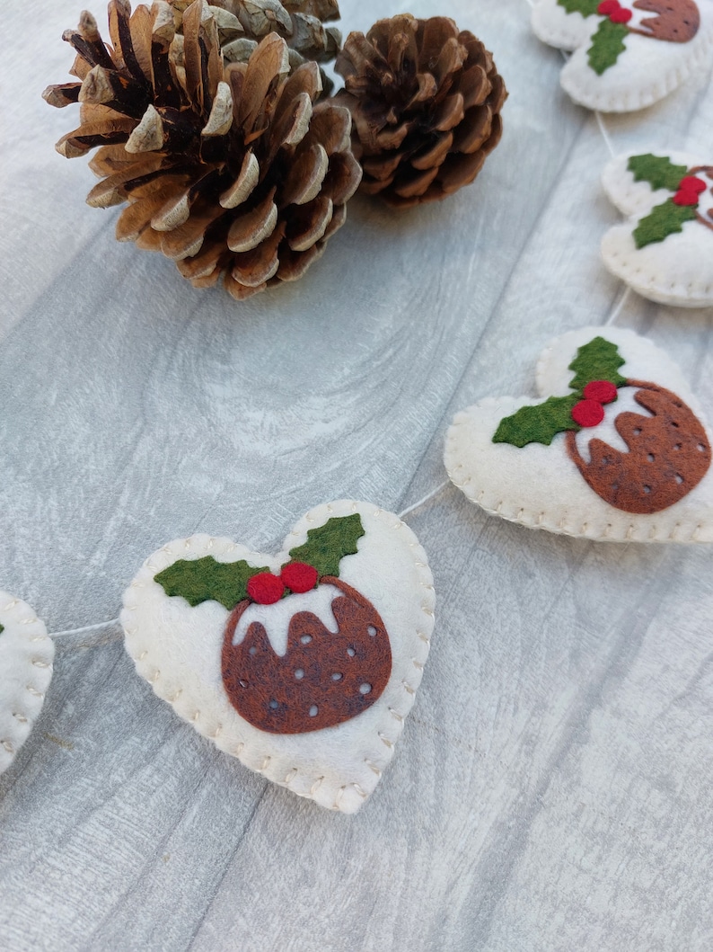 May include: A white felt Christmas garland with red and green felt Christmas pudding ornaments. The garland is strung with white thread and has a pine cone in the background.