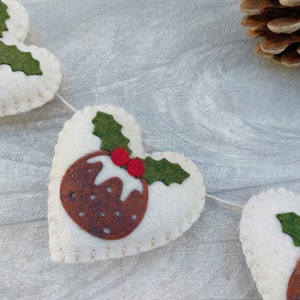 May include: A white felt Christmas garland with brown and green felt Christmas pudding ornaments. The garland is strung with white thread and has a white background.