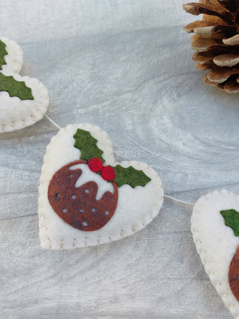 May include: A white felt Christmas garland with brown and green felt Christmas pudding ornaments. The garland is strung with white thread and has a white background.