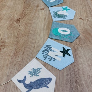 May include: A felt banner with the word "ONE" spelled out in white letters. Each letter is decorated with a different sea creature, including a seahorse, a turtle, a jellyfish, a starfish, and a whale. The banner is strung together with white twine.