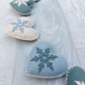 May include: A garland of felt heart-shaped decorations in shades of blue and cream, each featuring a snowflake design. The hearts are stitched together with white thread, creating a festive, winter-themed decoration.