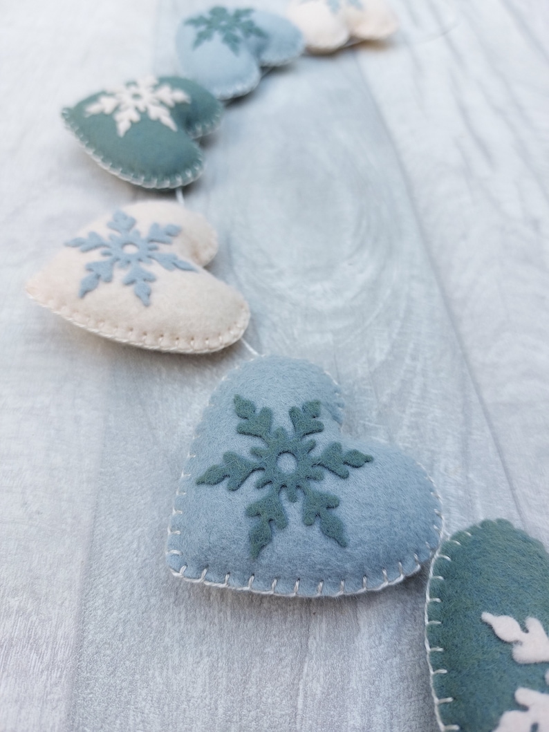 May include: A garland of felt heart-shaped decorations in shades of blue and cream, each featuring a snowflake design. The hearts are stitched together with white thread, creating a festive, winter-themed decoration.