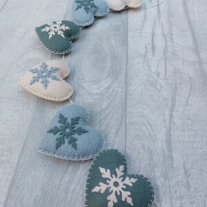 May include: A decorative garland featuring felt hearts in various shades of blue and cream, each embellished with a snowflake motif. The hearts are stitched with white thread and strung on a white cord, ideal for winter decor.