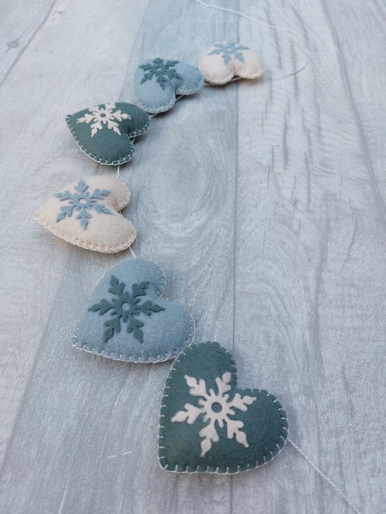 May include: A decorative garland featuring felt hearts in various shades of blue and cream, each embellished with a snowflake motif. The hearts are stitched with white thread and strung on a white cord, ideal for winter decor.