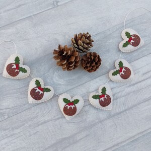 May include: A festive garland with white felt hearts decorated with brown felt Christmas pudding designs and green felt holly leaves. The garland is strung with white twine and is hanging against a light gray wood background.