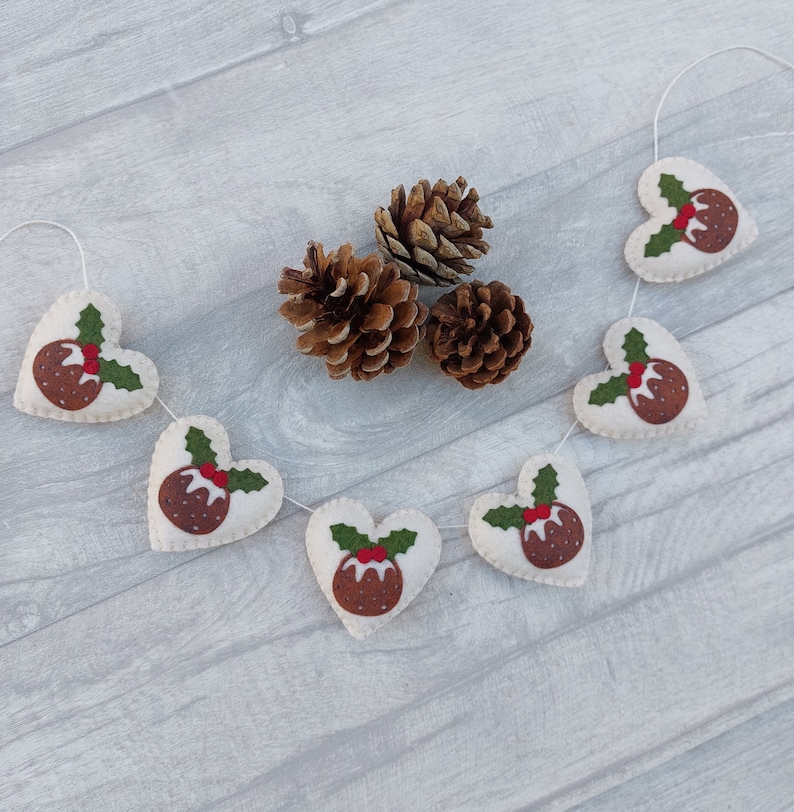 May include: A festive garland with white felt hearts decorated with brown felt Christmas pudding designs and green felt holly leaves. The garland is strung with white twine and is hanging against a light gray wood background.