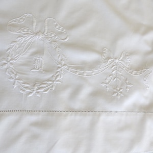 May include: A white textile featuring intricate white-on-white embroidery. The design includes a central wreath tied with a bow, enclosing the letter D, connected by a ribbon garland to a secondary bow and floral motif, with a hemstitch border.