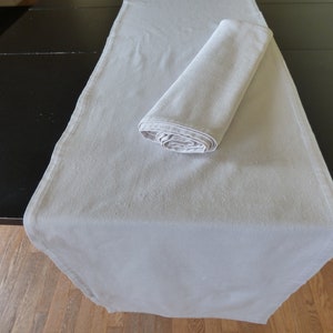 May include: A light gray table runner is rolled up and placed on a gray table runner. The table runner is made of a soft, textured fabric.