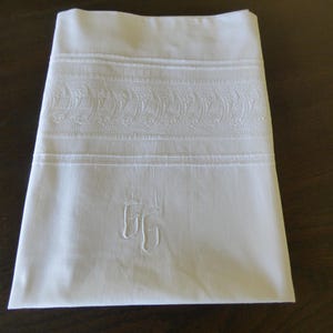 May include: A folded white cotton pillowcase featuring a wide horizontal band of intricate tone-on-tone floral embroidery and tuck stitching. The lower section displays a hand-stitched monogram with the letters GG in a matching white thread.