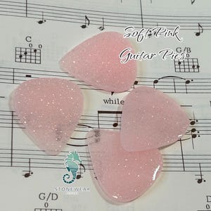 May include: Four soft pink guitar picks with a glittery finish. The picks are on a sheet of music with the text "Soft Pink Guitar Picks" written on it.