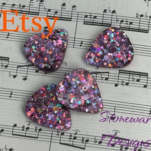 May include: Four purple and pink glitter guitar picks on a sheet of music. The picks are shaped like a triangle with rounded edges. The music sheet has the word "Etsy" printed at the top and "Stoneware Designs" printed at the bottom.