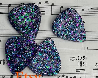 Könnte beinhalten: Vier Gitarrenplektren aus Harz mit eingebettetem blauem, grünem und violettem Glitzer. Die Plektronen sind dreieckig mit abgerundeter Spitze. Die Plektronen liegen auf einem Notenblatt mit dem Text "E9 Stonewear Designs A(add9)" und "Etsy" sichtbar.