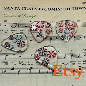 May include: Four clear guitar picks with red, white, and green peppermint candy designs. The picks are arranged on sheet music with the text "Santa Claus Is Comin' To Town" and the Etsy logo.