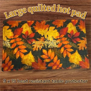 May include: Large, rectangular quilted hot pad with a fall leaf pattern in shades of yellow, orange, and red on a dark green background. The text "Autumn on The Table" is embroidered on the pad. Measures 9 x 15 inches.