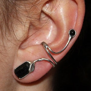 May include: A silver ear cuff with a black gemstone. The cuff wraps around the upper part of the ear and has a small black gemstone at the end.