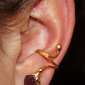 Könnte beinhalten: Ein goldener Ohrring mit einem herabhängenden violetten Edelstein. Der Ohrring umschließt das Ohr und hat eine kleine Kugel am Ende.