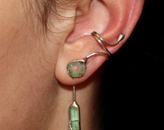 May include: A pair of earrings featuring a green gemstone and a silver ear cuff. The ear cuff is a twisted design that wraps around the ear. The gemstone earring is a long, thin, green stone.