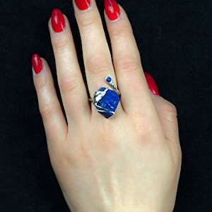 May include: A silver ring featuring a blue gemstone in the shape of a heart. The ring is worn on a finger with red nail polish.
