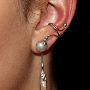 May include: A silver ear cuff with a pearl-like drop earring. The ear cuff wraps around the upper ear and has a small pearl-like accent. The drop earring features a long, pear-shaped pearl.
