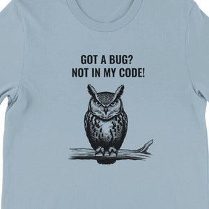 Könnte beinhalten: Hellblaues T-Shirt mit einer schwarz-weißen Illustration einer Eule auf einem Ast. Der Text "GOT A BUG? NOT IN MY CODE!" ist auf dem T-Shirt aufgedruckt.