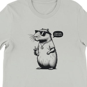 Puede incluir: Una camiseta gris claro con una ilustración en blanco y negro de un roedor de dibujos animados con gafas de sol y sosteniendo una bebida. El roedor tiene un globo de diálogo que dice "Código, dormir, repetir."