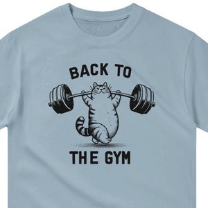 Könnte beinhalten: Hellblaues T-Shirt mit einer schwarz-weißen Illustration einer Katze, die eine Langhantel hebt. Der Text "BACK TO THE GYM" ist auf dem T-Shirt aufgedruckt.