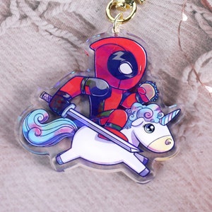 May include: A clear acrylic keychain featuring a cartoon Deadpool character riding a white unicorn. The keychain has a gold star-shaped clasp. Deadpool is in his red and black suit, holding a sword.
