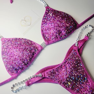 May include: A vibrant magenta bikini set, embellished with sparkling rhinestones. The top features triangle cups with rhinestone accents, and the bottoms have a matching design. The bikini is designed for competition wear.