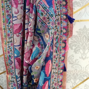 Puede incluir: Bufanda colorida con estampado floral en tonos rosa, azul y verde. La bufanda tiene un borde con un dise&ntilde;o repetitivo y est&aacute; rematada con borlas. Colgada de una percha negra.