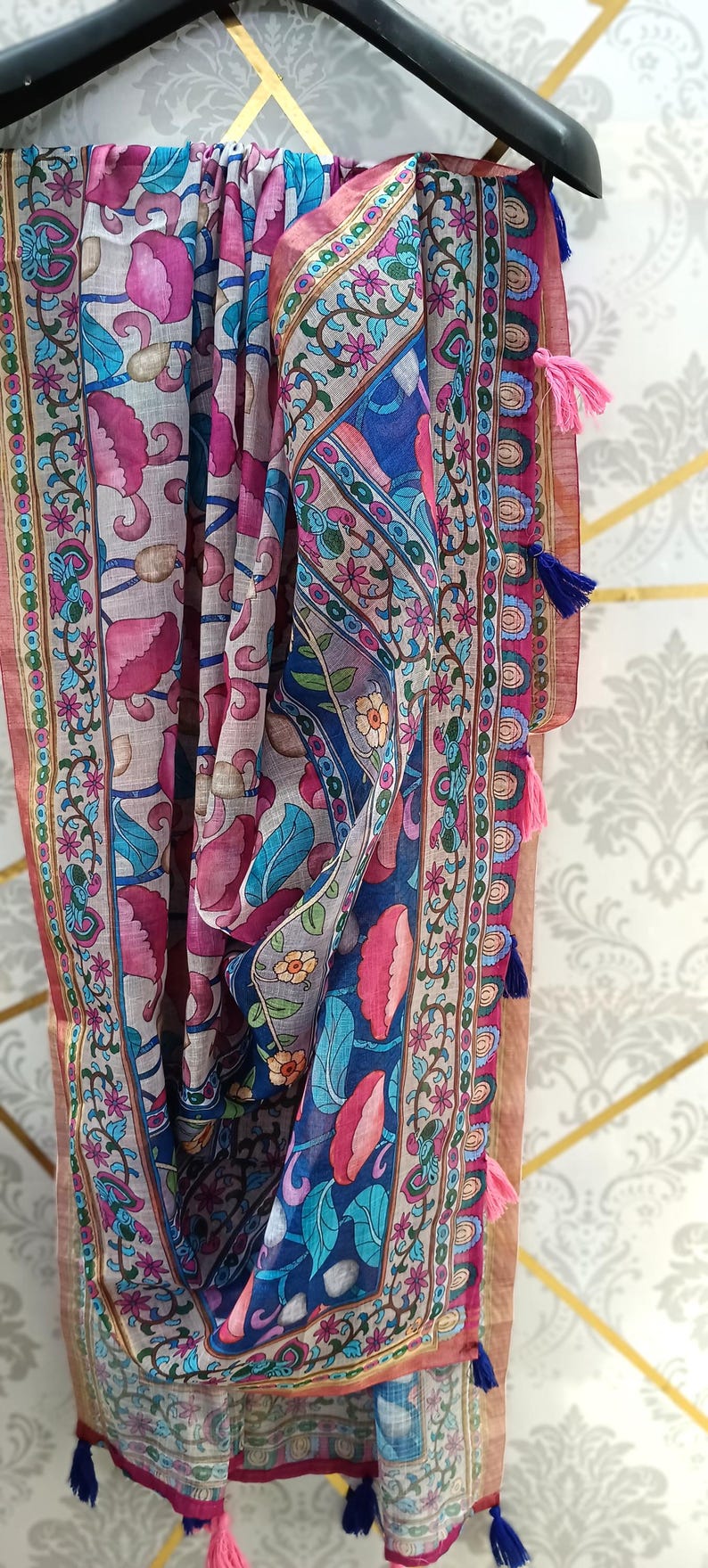 Puede incluir: Bufanda colorida con estampado floral en tonos rosa, azul y verde. La bufanda tiene un borde con un dise&ntilde;o repetitivo y est&aacute; rematada con borlas. Colgada de una percha negra.