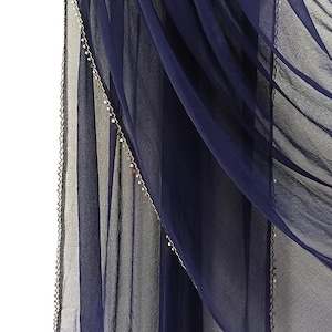 May include: A navy blue sheer scarf is draped over a black hanger. The scarf has a delicate beaded trim along the edges. The fabric is lightweight and flowing, creating soft folds.