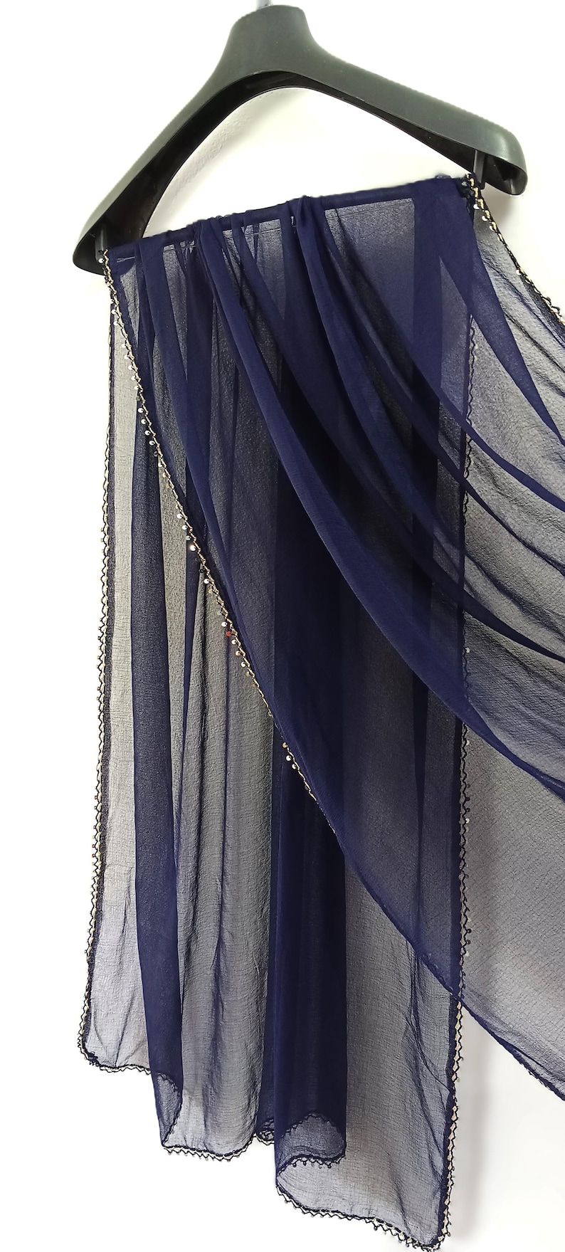 May include: A navy blue sheer scarf is draped over a black hanger. The scarf has a delicate beaded trim along the edges. The fabric is lightweight and flowing, creating soft folds.