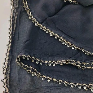 May include: A dark navy blue scarf with a delicate, scalloped edge. The edge is embellished with small, shiny, silver-colored sequins and a gold-colored trim. The fabric appears to be lightweight and flowing, with subtle folds and creases.