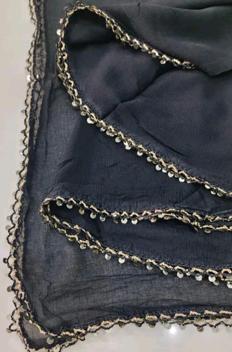 May include: A dark navy blue scarf with a delicate, scalloped edge. The edge is embellished with small, shiny, silver-colored sequins and a gold-colored trim. The fabric appears to be lightweight and flowing, with subtle folds and creases.