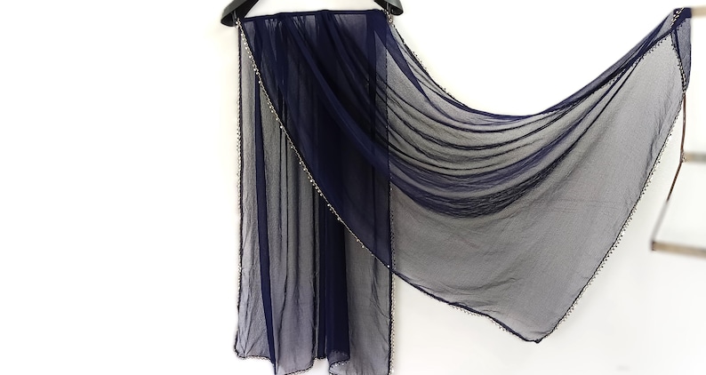 May include: A sheer navy blue scarf with a delicate beaded trim along the edges. The scarf is draped over a black hanger, showcasing its flowing, lightweight fabric. The scarf's design is simple and elegant.