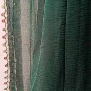 May include: Close-up of a dark green fabric with a textured appearance. The fabric is draped, creating soft folds and shadows. A decorative trim with colorful beads and a scalloped edge adorns the left side, adding a touch of detail.