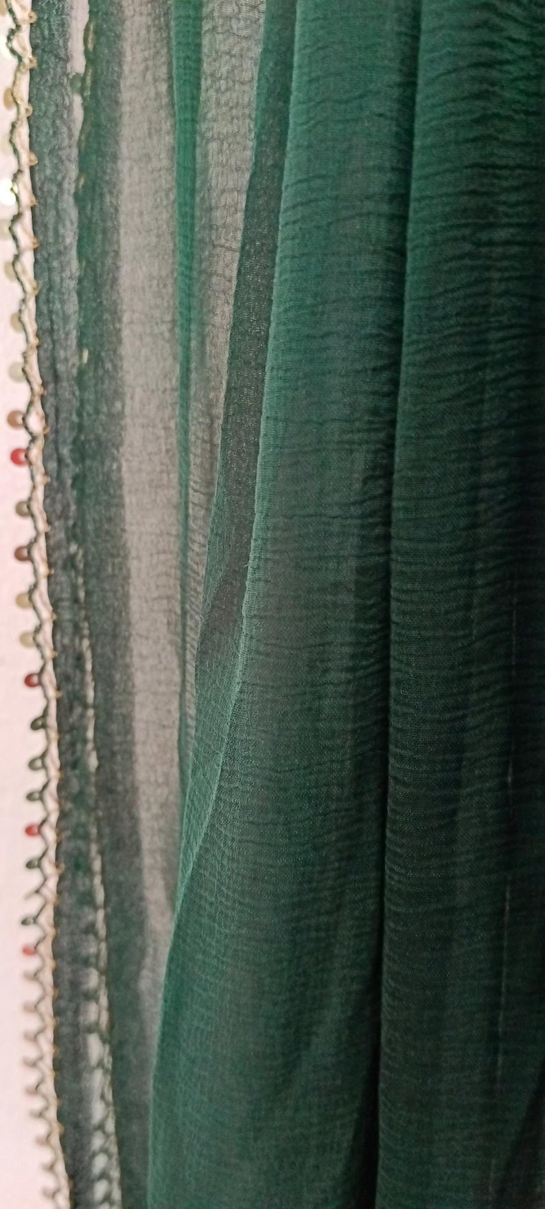 May include: Close-up of a dark green fabric with a textured appearance. The fabric is draped, creating soft folds and shadows. A decorative trim with colorful beads and a scalloped edge adorns the left side, adding a touch of detail.