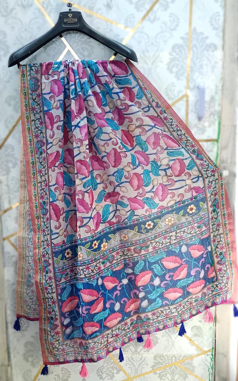 Puede incluir: Bufanda estampada con motivos florales en tonos rosa, azul y beige. El dise&ntilde;o presenta flores y hojas estilizadas, con un borde azul oscuro y borlas en los extremos. La bufanda est&aacute; colgada de una percha negra.