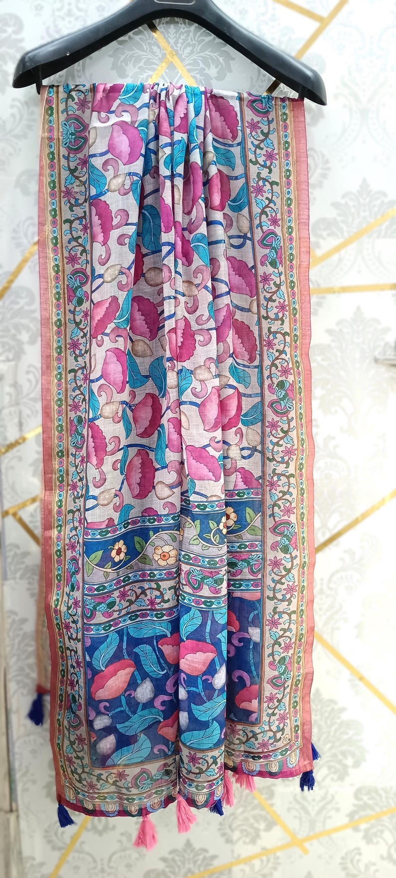 Puede incluir: Bufanda estampada con motivos florales rosas y azules sobre fondo claro. Tiene un borde azul y borlas rosas. Se muestra en una percha negra, mostrando su dise&ntilde;o.