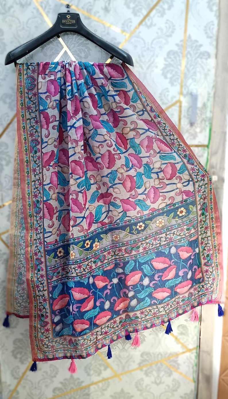 Puede incluir: Bufanda estampada con motivos florales en tonos rosa, azul y beige. El dise&ntilde;o presenta flores y hojas estilizadas, con un borde decorativo y borlas. La bufanda est&aacute; colgada de una percha negra.