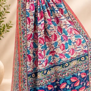 Puede incluir: Bufanda estampada con motivos florales en rosa, azul y blanco. La bufanda presenta un dise&ntilde;o repetitivo de flores rosas y hojas azules sobre un fondo blanco. Los bordes est&aacute;n adornados con un borde decorativo y borlas.