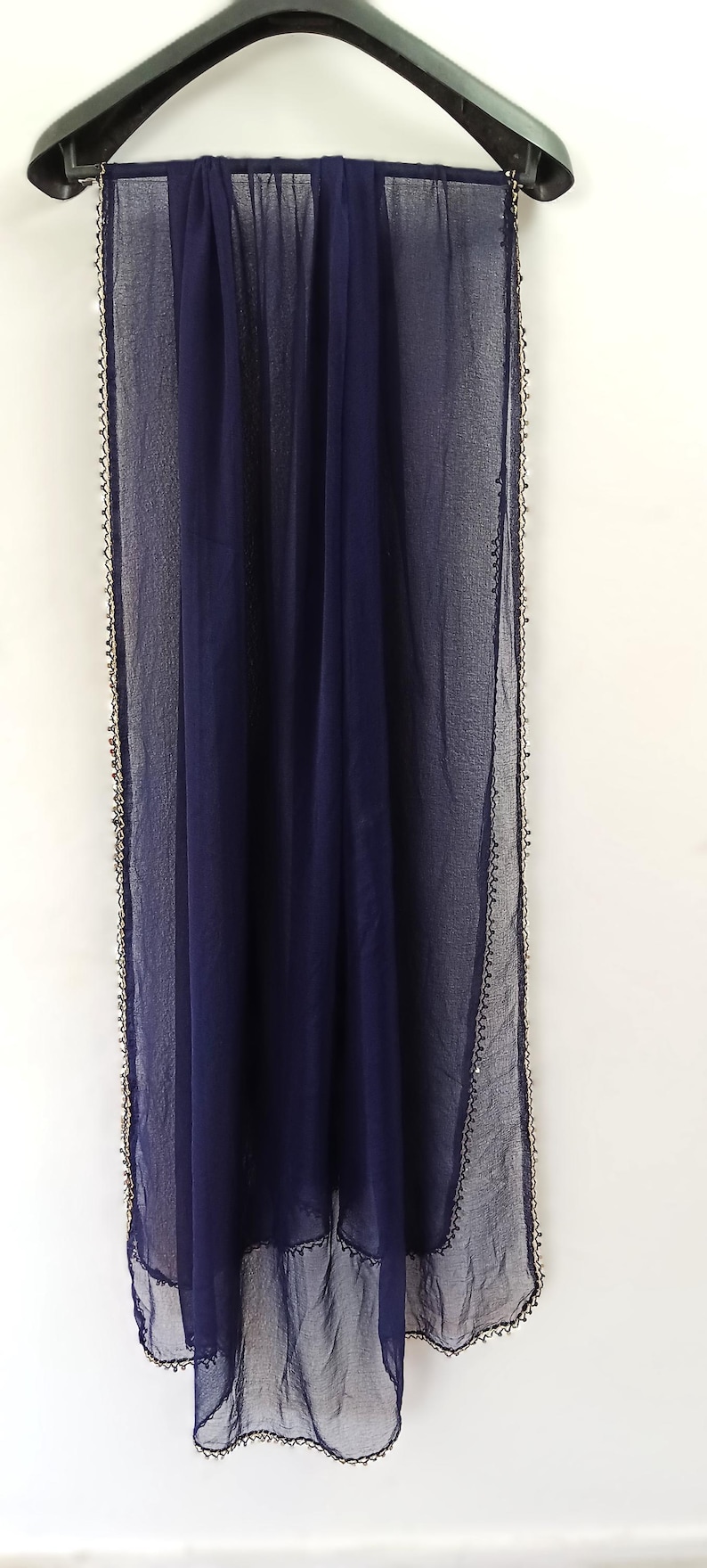 May include: A navy blue sheer scarf with a delicate, beaded trim. The scarf is hanging on a black hanger against a white background. The fabric appears lightweight and flowing, suitable for various occasions.