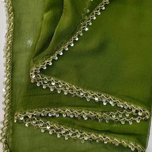 May include: Olive green fabric with a delicate, shimmering trim. The trim is a light gold color and is adorned with small, round sequins. The fabric is draped, creating soft folds and shadows. The overall effect is elegant and refined.