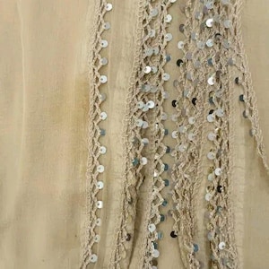 May include: Close-up of a beige fabric with decorative trim. The trim features a crocheted design with silver sequins. The fabric appears to be lightweight and flowing, with a slightly wrinkled texture. The overall aesthetic is delicate and elegant.