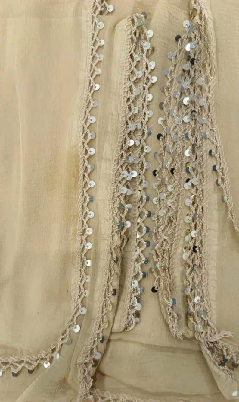 May include: Close-up of a beige fabric with decorative trim. The trim features a crocheted design with silver sequins. The fabric appears to be lightweight and flowing, with a slightly wrinkled texture. The overall aesthetic is delicate and elegant.