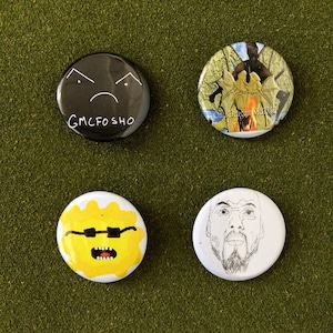 May include: Four round button pins with different designs. The first pin is black with a white frowning face and the text "GMCFOSHO". The second pin has a colorful image of a dragon with a fire in front of it. The third pin is yellow with a pair of sunglasses and a mouth open in a scream. The fourth pin is white with a black and white drawing of a man's face.