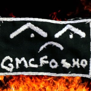 May include: A black fabric patch with white stitching. The patch features a sad face with two eyebrows and a frown. The text "GMCFO SHO" is written below the face.