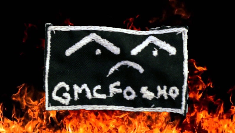 Puede incluir: Un parche de tela negro con costuras blancas. El parche presenta una cara triste con dos cejas y un ce&ntilde;o fruncido. El texto "GMCFO SHO" est&aacute; escrito debajo de la cara.