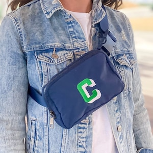 Nylon Navy Belt bag, worn across the chest of a model in a denim jacket. Fanny pack is embroidered with a Kelly green block "C" with a white shadow.