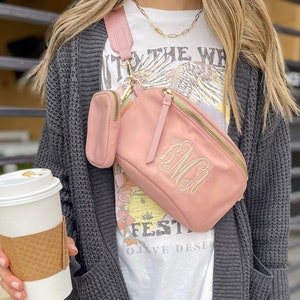 Blush colored, nylon sling bag across the chest of a model holding coffee. Model is also wearing a t-shirt and cardigan. Monogrammed letters in Ivory thread decorate the front of the crossbody. A webbed, adjustable strap is the same hue as the bag.